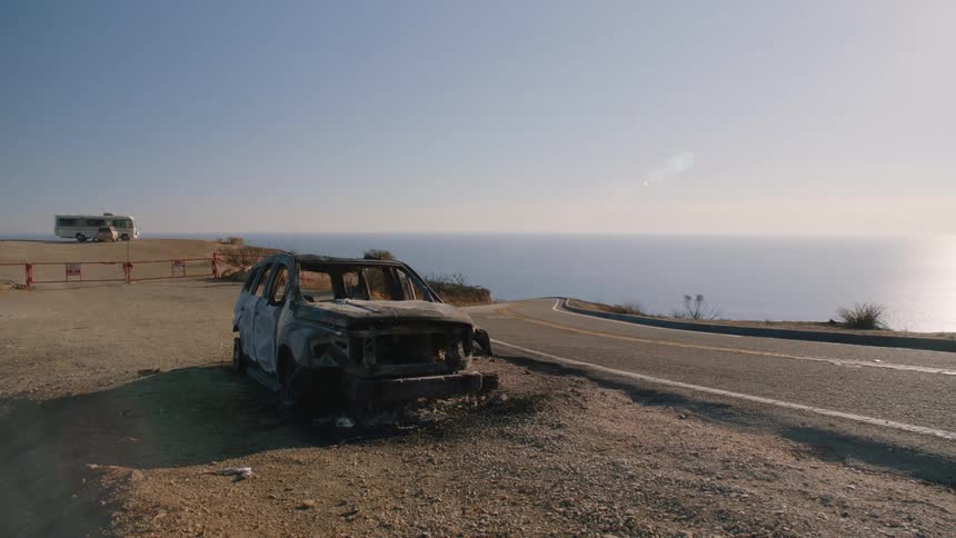 An old abandoned car on the side of the road and a motor home parked in the distance witht he ocean on the horizon