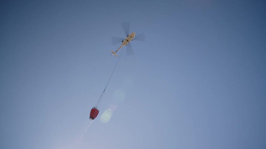 A helicopter carries a specialized bucked suspended on a cable to deliver water for aerial firefighting
