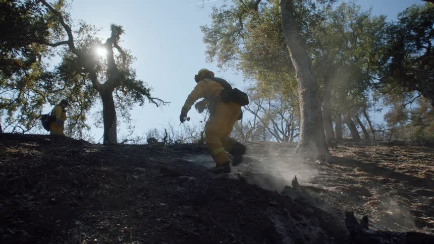A firefighter carries an axe as he walks up a hill near the woods as smoke rises in the air