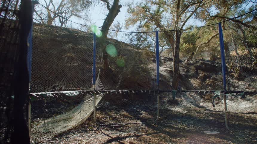 A view of a ruined hammock and trampoline cage near a dirt hill and sparse trees