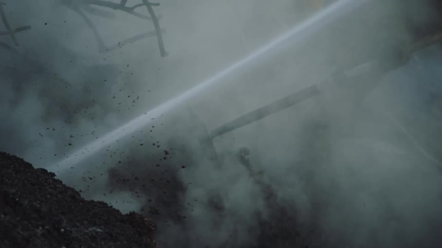 A fireman is raking the soil while a firehose is soaking the area with water as they fight a smoldering area in a forest fire