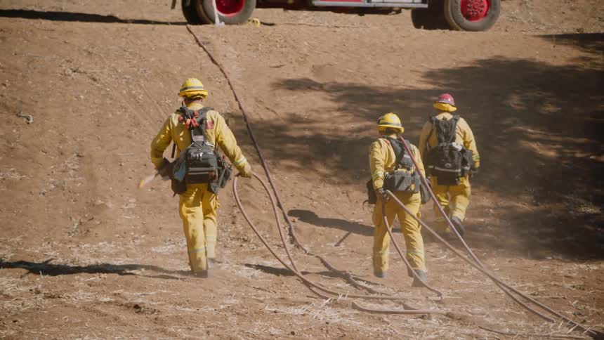Three firefighters are dragging a hose up a dirt hill to the fire truck