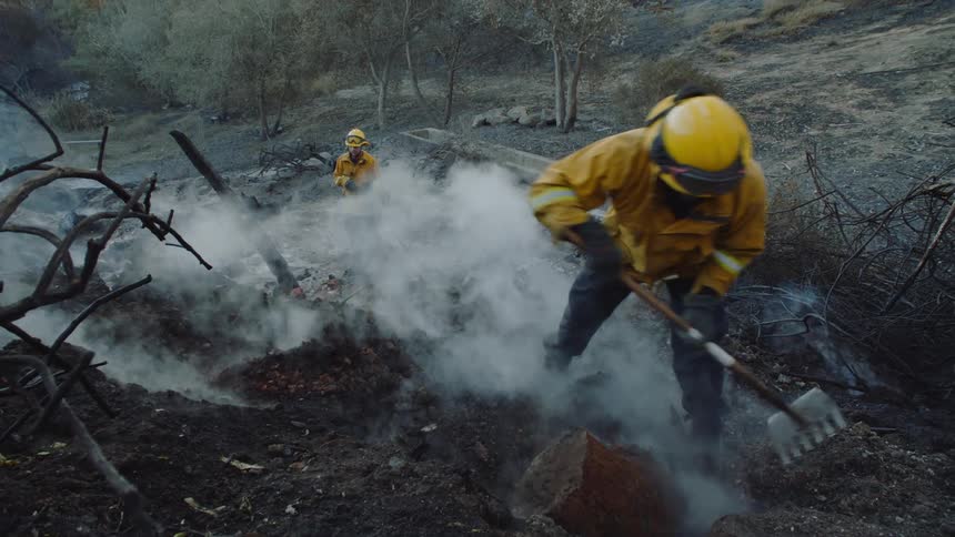 Firefighters dig in dirt and move rocks as smoke billows in a forest