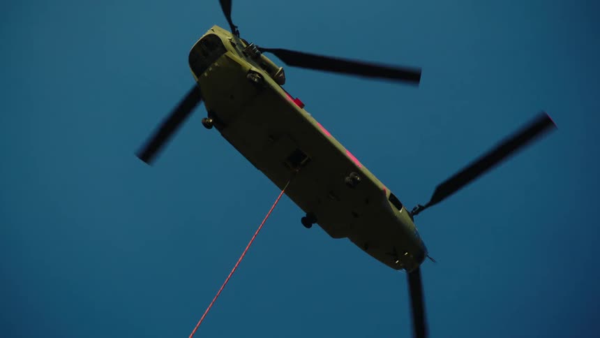 A large green helicopter hovering with a cable descending from the helicopter