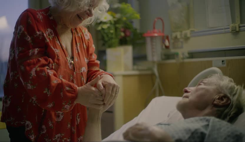 A elderly woman coming to sit on the hospital bed of an elderly man while clasping his hand