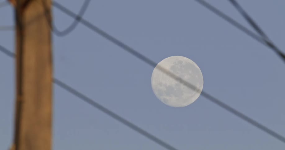 The full moon shines through utility wires in a clear blue sky