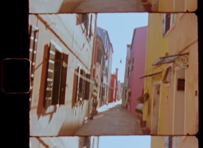 Shaky POV film footage walking through a narrow alley or street with old and colorful houses and buildings with windows and entrance doors as the sun shines and it covers the alleyway in shadow