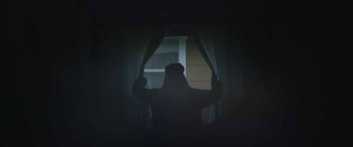 A woman walks into a dark room stops by the windows turns to the left and opens the drapes and then takes a look out the window watching the neighboring building