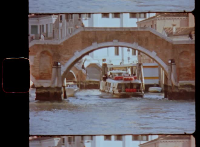 A large motor boat or ferry drives or moves under a pedestrian bridge in waterway in an old city or town build next to the water on vintage Super 8 film footage