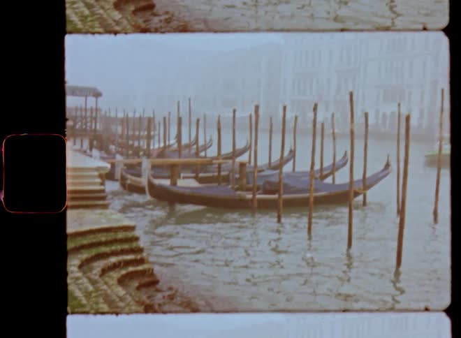 Long canoes or Venetian gondola sit parked in a row on a pier tied up to wooden poles moving in the waves as another boat drives by on the water of the canals on a foggy spring time morning
