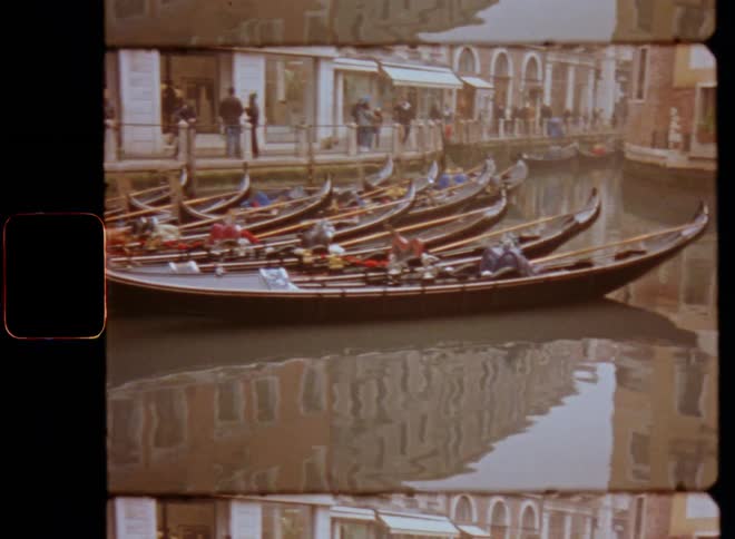Several boats or gondolas parked next to each other in a harbor or port in the Venice canals as people or tourists are walking on a pier or jetty in front of shops caught on vintage film