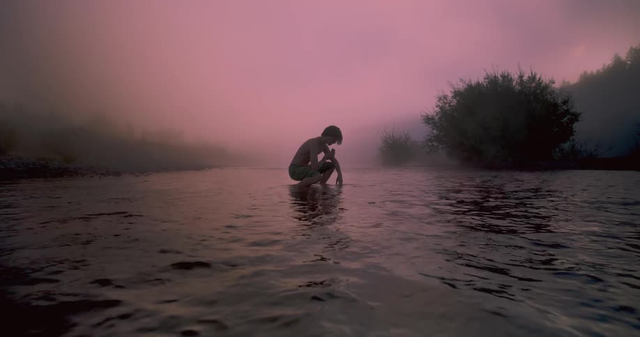 A male squats in a lake and runs his hand through the water