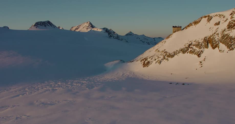 An aerial of snow covered mountains and plateaus with a building on the mountain ledge