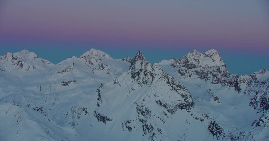 Aerial twisting footage of the snow covered slopes of a mountain range under a colorful evening sky
