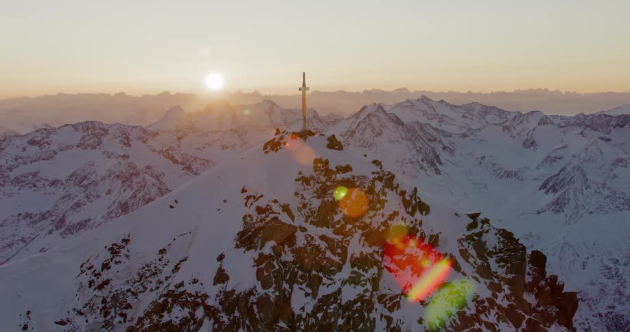 A cross stands on a snow covered mountain peak as the sun shines brightly on the horizon and there are mountains in the distance
