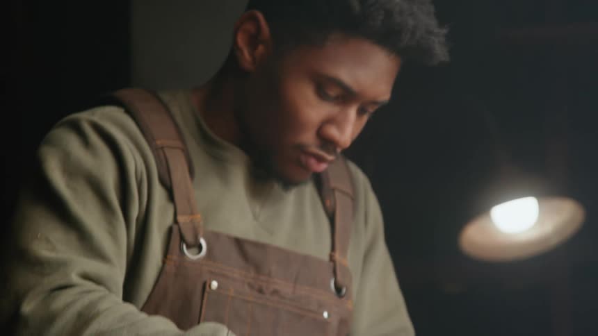 A man is standing next to a table looking down as he is woodworking with a small piece of wood and a chisel