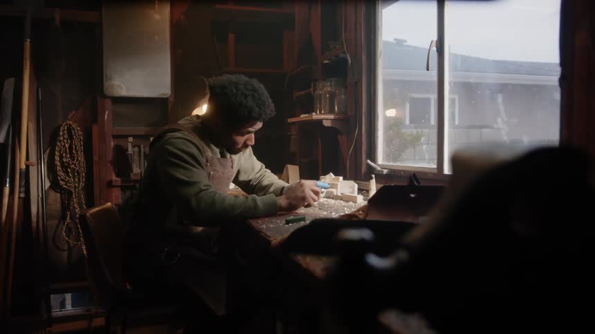 Woodcarver sitting at workbench near window carves wood