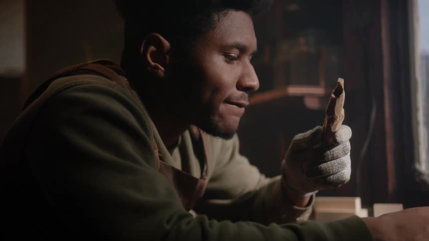 A man is sitting at a table near a window looking at small piece of carved wood