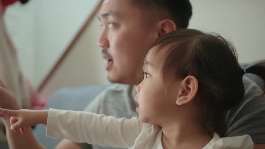 A toddler girl sitting on her fathers lap as they point and look away from them