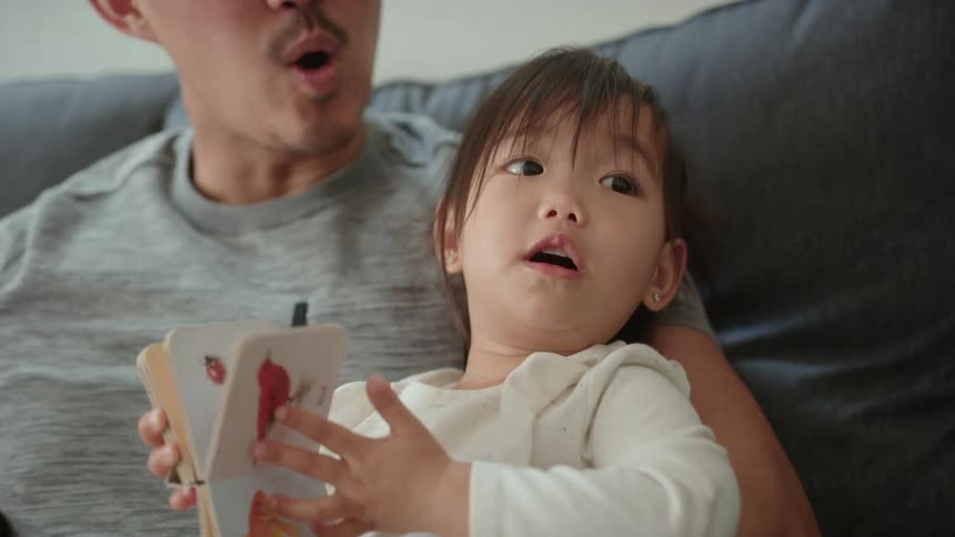 A father and his young daughter wave to someone while sitting on the sofa
