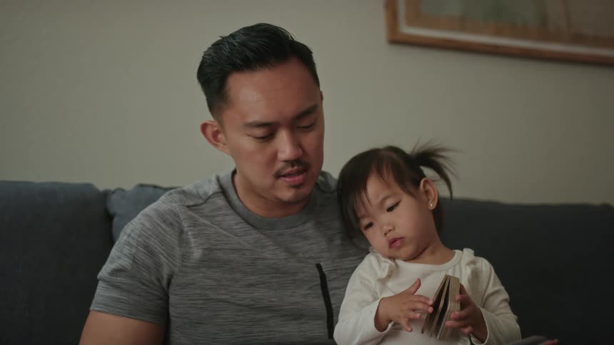 A father reads a book to his daughter on the couch while she sits on his lap
