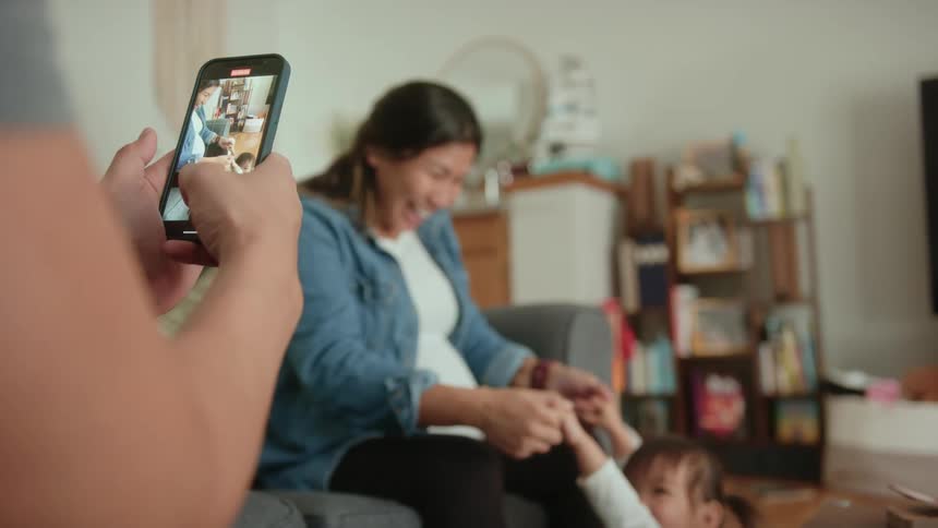 Father uses his smartphone to take video of his pregnant wife and daughter playing