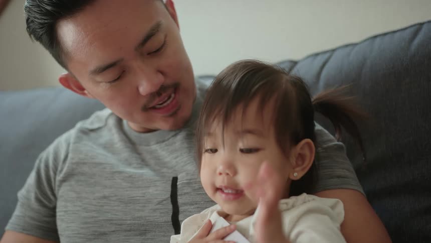 A father holding his little girl while he reads to her on the couch and they express happiness