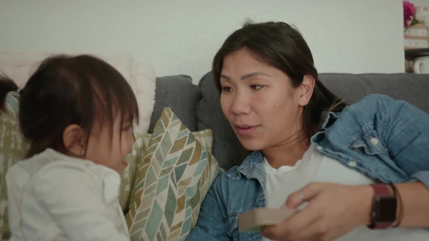 Mother with denim jacket and white shirt plays with toddler girl sitting on gray couch in the living room