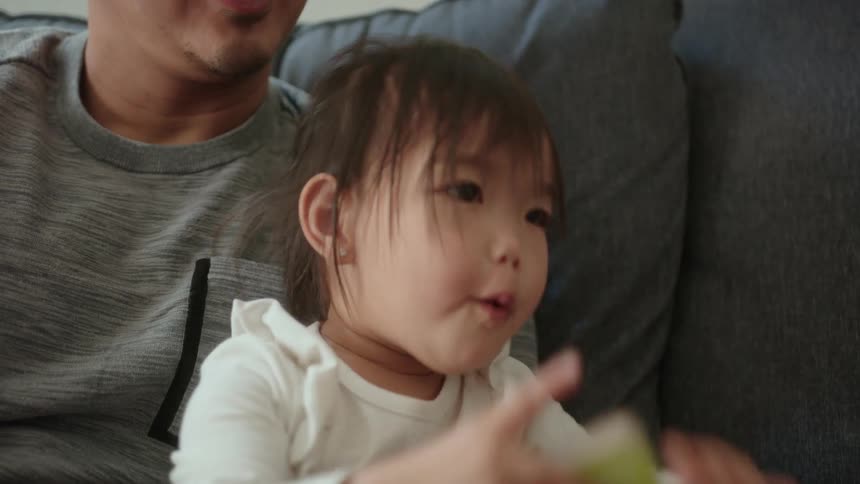 A little girl is sitting in her fathers lap as she holds a small book and crawls across the couch