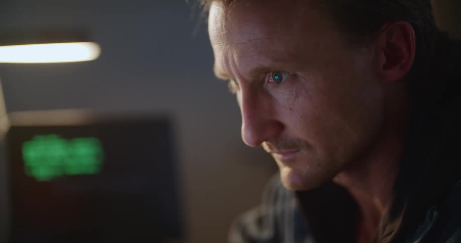 A man sits at his desk while lines of code process on a computer beside him