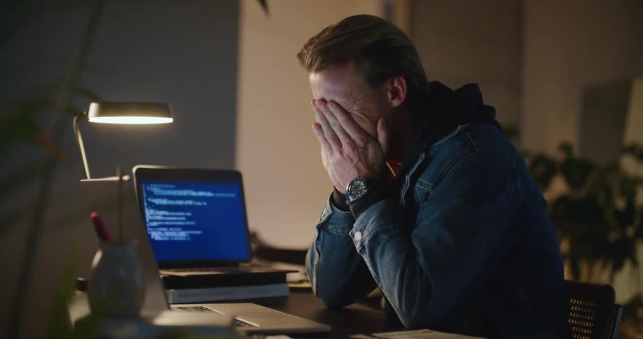 A man is sitting at a desk typing on a laptop computer and looking over at another screen as he types and he stops to think