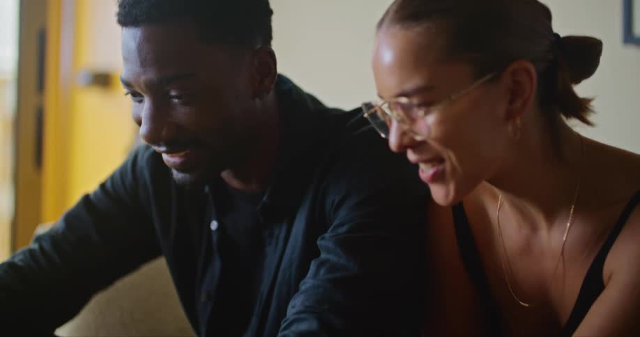 Man sits on couch smiling while man manipulates laptop using touch pad
