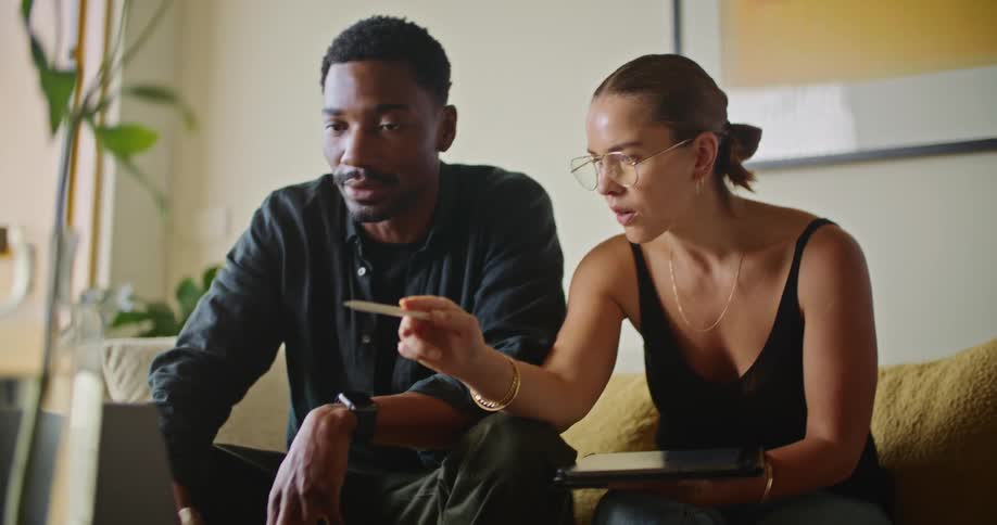 A man and a woman are sitting on the living room couch talking as they look at a laptop computer and she is holding a pen pointing to the screen