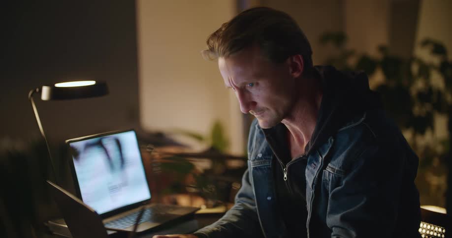 A man sitting at the computer desk reads some notes he took of a piece of paper glances at the laptop screen while thinking and then looks at an open book sitting on the desk