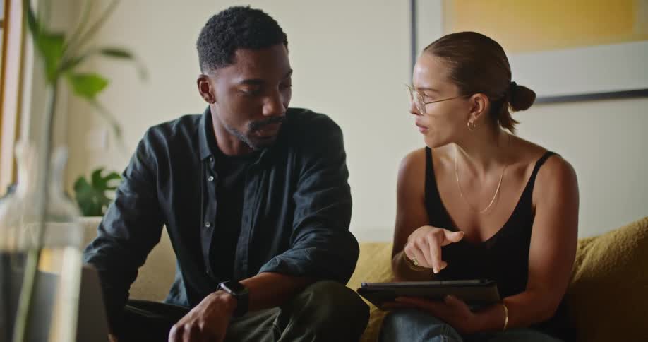 A woman sits on a couch holding a tablet next to a man working on a laptop as they talk with one another