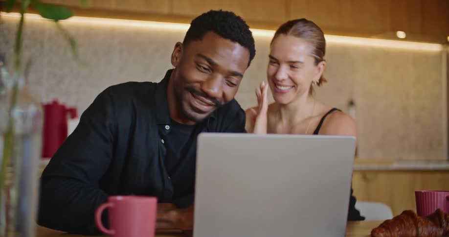 A young couple are sitting at the kitchen table watching a laptop screen looking happy and the woman kisses and hugs the man while smiling as if they are celebrating something