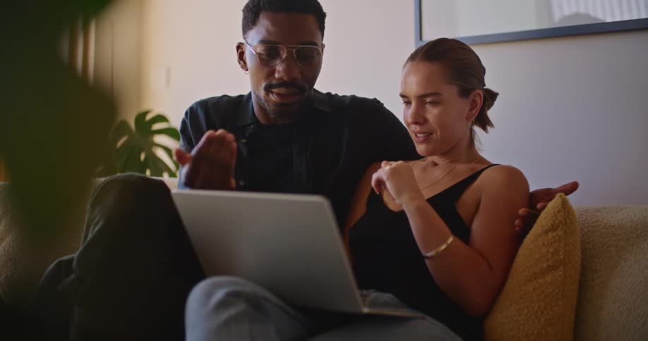A man and woman sitting on a couch with a laptop and pointing at it
