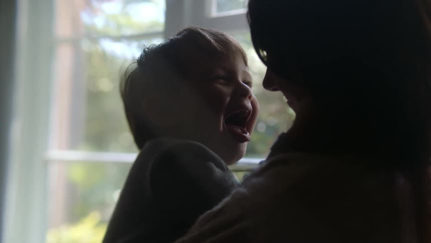 A boy in his mothers arms in front of a window as the boy laughs and makes face at his mother
