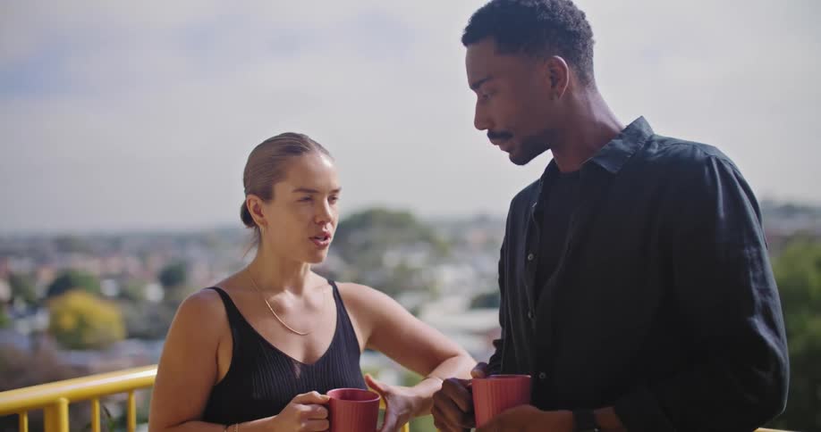 A woman and man stand outdoors while conversing as they hold coffee mugs