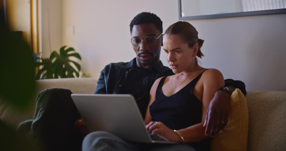 A man and a woman are sitting on the couch talking as they look at a laptop computer and he has his arm around her