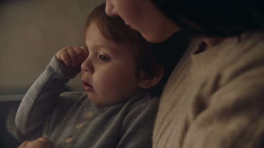 A woman is holding her young son in her lap and talking to him as he rubs his eyes with his hand