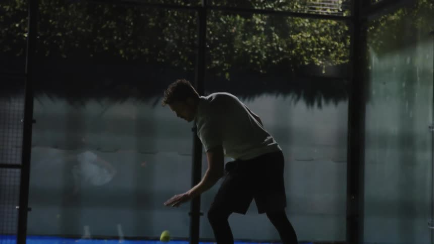 A man standing on a tennis court at night uses a racket to serve and hit a ball