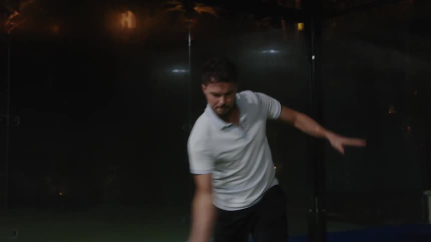 A man outdoors at night uses a tennis racket to hit an underhand serve ball