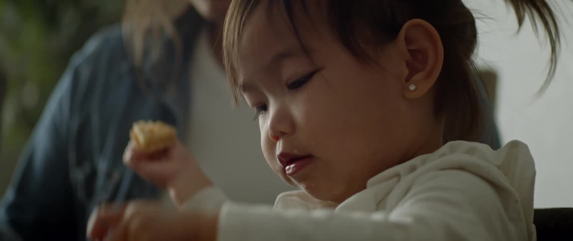 A little girl is sitting with her mother as she eats a cookie she is holding in her hand