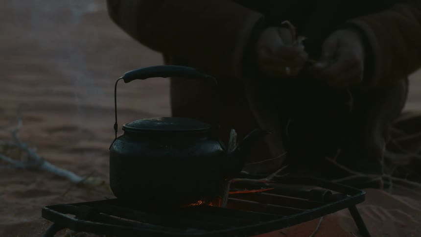 Hand of a person stirring the contents of a teapot over a fire in the desert sands at evening