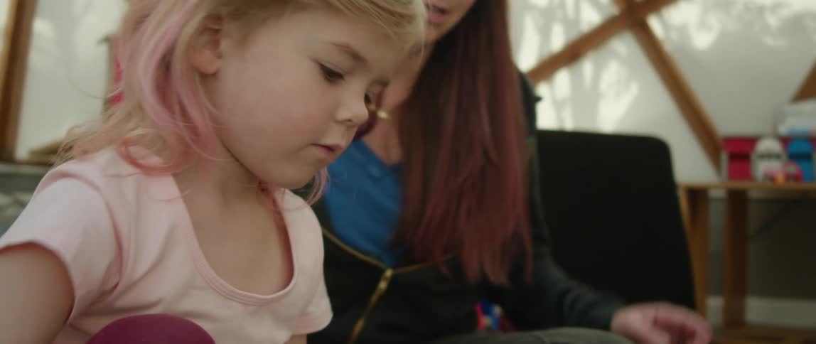 Girl with her hair tinted pink sit on the floor of a room playing with toys at daytime with her mother