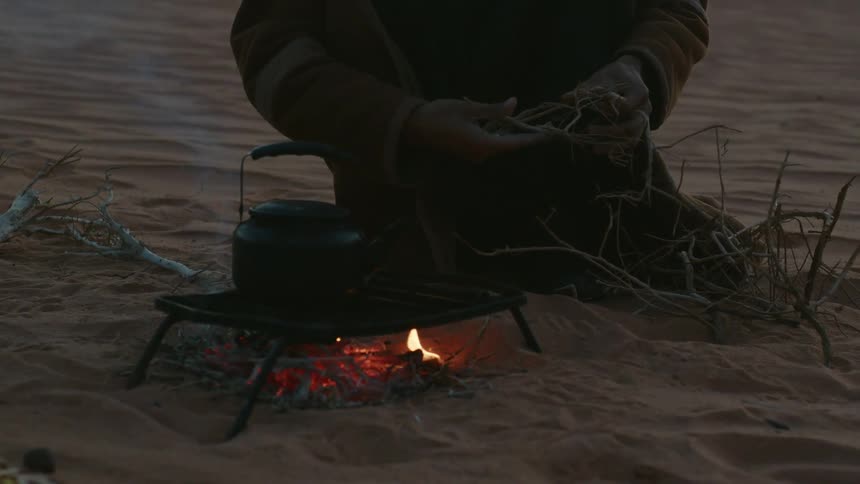Hand of a person breaking dry roots over a grill next to a teapot in the desert
