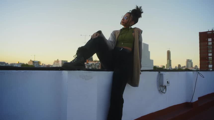 A young woman reclines on the edge of a building while gazing out over an urban skyline in contemplation