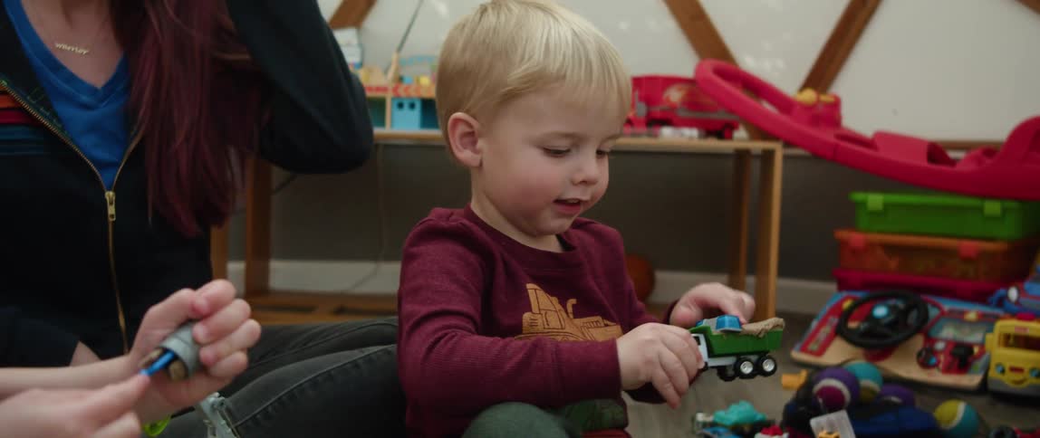A little boy is sitting on a playroom floor surrounded by toys as he plays with trucks in a sandbox