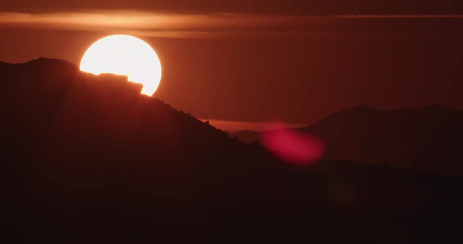 A big bright sun sets behind the silhouette of a mountain in a deep red sky in the desert